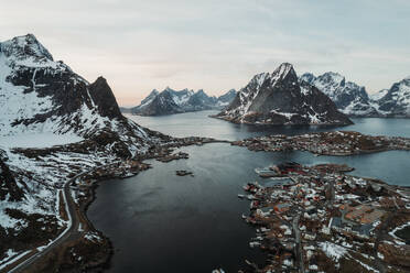 Scenic view of massive rocky mountains with snowy and frozen peaks by residential onshore settlement over river in winter at daylight - ADSF46609