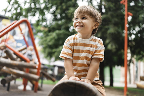 Lächelnder Junge sitzt auf einem Spielgerät im Park - ANAF01935