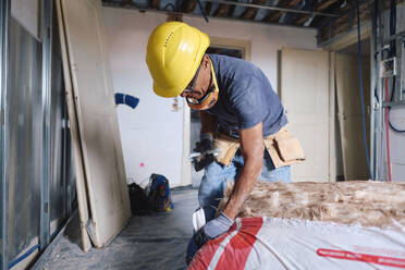 Älterer Bauarbeiter mit Schutzhelm, der auf einer Baustelle mit Isolierung arbeitet - ASGF04292