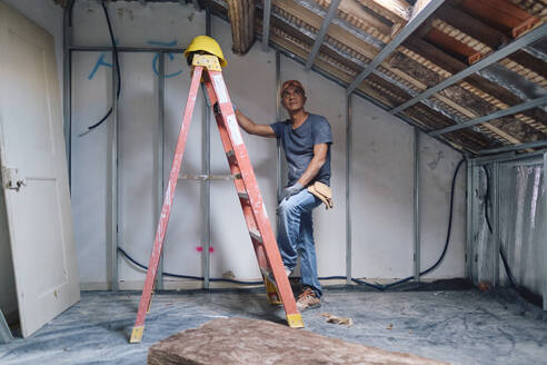 Nachdenklicher reifer Bauarbeiter, der auf der Baustelle auf einer Leiter steht - ASGF04302