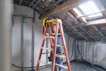 Bauarbeiter mit Schutzhelm, der auf der Baustelle hinter einer Leiter steht - ASGF04303