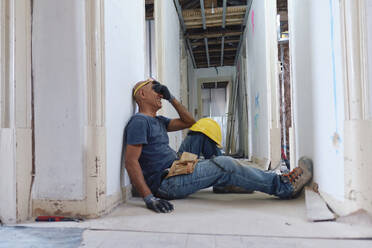 Happy mature construction worker covering eyes with hand sitting in corridor - ASGF04318