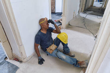 Tired construction worker touching forehead and sitting in corridor - ASGF04319