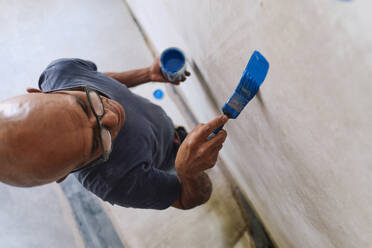 Glatzköpfiger Bauarbeiter, der blaue Farbe auf die Wand aufträgt - ASGF04340