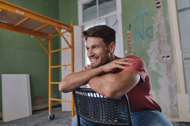 Happy man sitting on chair with arms crossed at site - ASGF04370