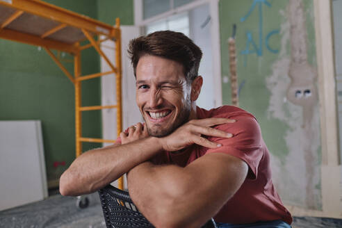 Happy man winking and sitting on chair at site - ASGF04371