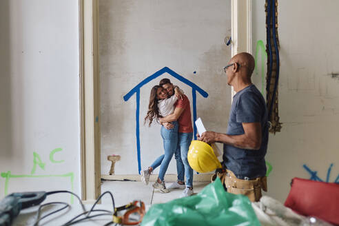 Bauarbeiter mit fröhlichem Paar, das sich auf der Baustelle umarmt - ASGF04391