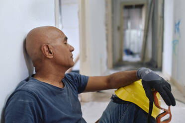 Tired construction worker sitting with eyes closed in corridor - ASGF04404