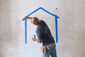 Glatzköpfiger Bauarbeiter, der auf der Baustelle mit einem Pinsel ein Haus an die Wand malt - ASGF04411