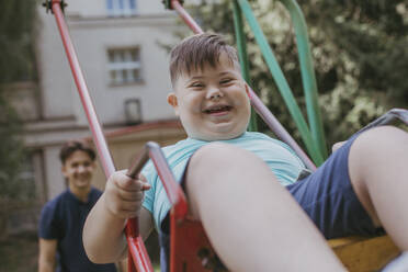 Glücklicher Junge mit Down-Syndrom spielt auf einer Schaukel mit seinem Bruder im Hintergrund - OSF01992