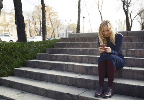 Blonde Frau mit Mobiltelefon auf einer Treppe sitzend - AZF00563