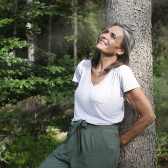 Smiling woman leaning on tree in forest - JBYF00232