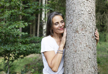 Smiling woman hugging tree in forest - JBYF00234