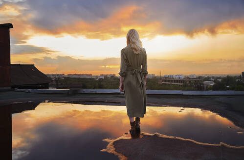 Blonde junge Frau steht an einer Pfütze auf einem Dach unter bewölktem Himmel - AZF00577