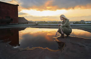 Blond woman touching water puddle on rooftop at sunset - AZF00578