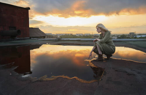 Blonde Frau berührt Wasserpfütze auf dem Dach bei Sonnenuntergang - AZF00578