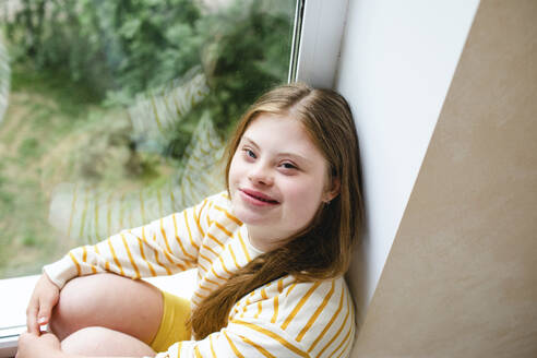 Smiling teenage girl with down syndrome sitting on window sill - MDOF01427