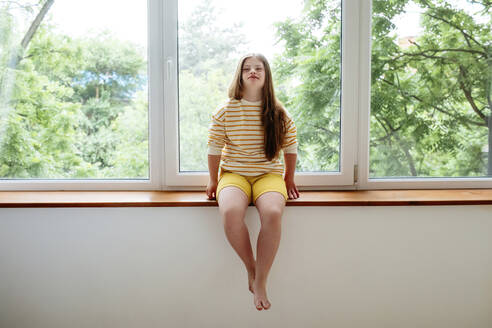 Teenager-Mädchen mit Down-Syndrom sitzt zu Hause auf der Fensterbank - MDOF01430