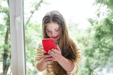 Mädchen mit Down-Syndrom benutzt ein Smartphone vor einem Fenster - MDOF01431