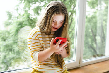 Mädchen mit Down-Syndrom benutzt ein Mobiltelefon auf der Fensterbank - MDOF01432