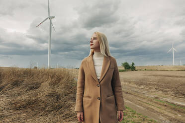 Blonde Frau steht vor einer Windkraftanlage unter bewölktem Himmel - VSNF01289