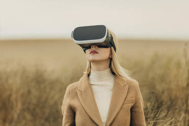 Blond woman wearing virtual reality simulators standing in field - VSNF01300