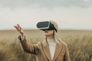 Blond woman gesturing with virtual reality simulators in front of field - VSNF01302