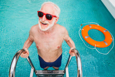 Happy senior man having party in the swimming pool - Active elderly male person sunbathing and relaxing in a private pool during summertime - DMDF02681