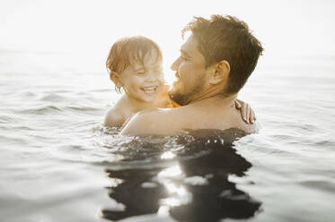 Fröhlicher Vater mit Sohn beim Schwimmen im Wasser an einem sonnigen Tag - ANAF01953