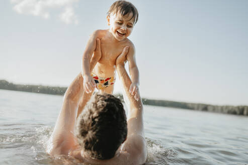 Cheerful son enjoying with father in water on sunny day - ANAF01954