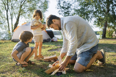 Vater und Kinder mit Holz für ein Lagerfeuer auf einer Wiese - ANAF01960