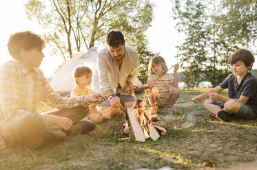 Vater und Kinder braten gemeinsam Würstchen am Lagerfeuer beim Picknick - ANAF01965