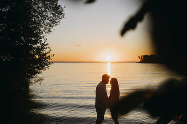 Mann umarmt Frau vor dem Meer bei Sonnenuntergang - ANAF01972
