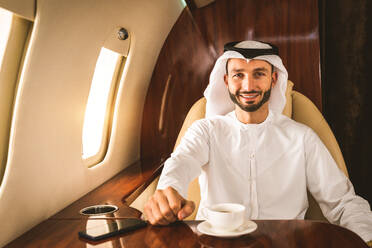 Arabischer Mann mit Kandora im Emirates-Stil fliegt in exklusivem Privatjet - Nahöstlicher Geschäftsmann mit traditioneller Kleidung fliegt in exklusiver Business Class im Flugzeug, Konzepte über Business und Transport - DMDF02768