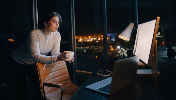 Thoughtful businesswoman works late at night in her home office, focused on a digital marketing project. Professional woman working with a computer and a cup of coffee, trying to meet a deadline. - JLPSF30731