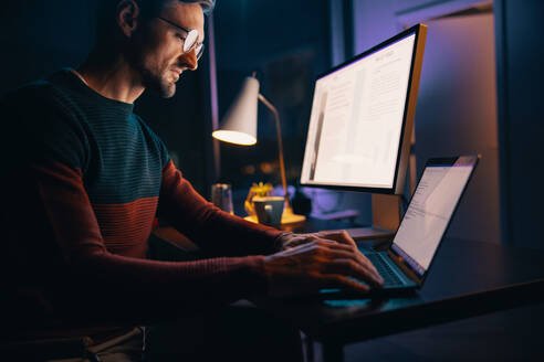 Kaukasischer Mann, der spät nachts in seinem Heimbüro an einem Projekt arbeitet und dabei einen Laptop benutzt. Geschäftsmann, der die Technologie nutzt, um nach Feierabend bei der Fernarbeit produktiv zu sein. - JLPSF30735