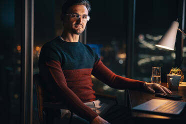 Dedicated businessman looking at the camera while sitting at his office desk after hours. Professional man working overtime to meet his project deadlines. - JLPSF30737