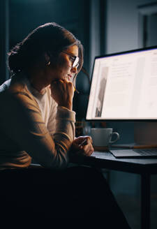 Pensive businesswoman works on her laptop in a home office late at night. Focused and determined, she plans and strategizes for her project, showing her dedication to the business. - JLPSF30739