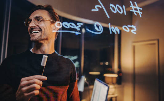 Ein glücklicher kaukasischer Geschäftsmann arbeitet bis spät in die Nacht in einem Büro mit Glaswänden und schreibt Notizen. Er ist ein kreativer Datenanalyst, Unternehmer und Datenwissenschaftler, der sich seinem Projekt widmet. - JLPSF30741