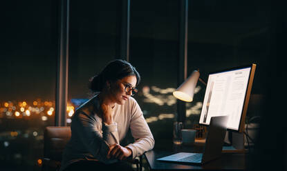 Eine Geschäftsfrau arbeitet noch spät in einem schwach beleuchteten Büro an einer virtuellen Besprechung. Sie sitzt an ihrem Schreibtisch und hört ihren Kollegen während einer nächtlichen Videokonferenz aufmerksam zu. - JLPSF30752