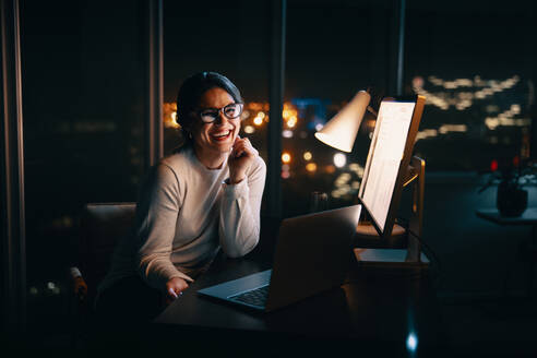 Glückliche Geschäftsfrau, die in die Kamera lächelt, während sie noch spät in ihrem Büro arbeitet. Professionelle Frau, die lange arbeitet, um ihre Produktivität zu steigern und ihre Projektfristen einzuhalten. - JLPSF30759