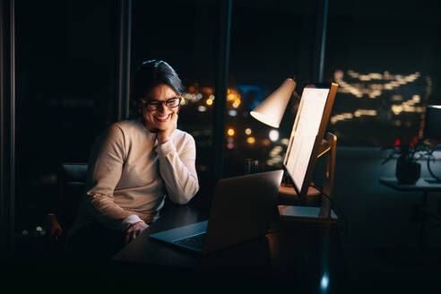 Businesswoman works late from her home office, engaging in a productive video call with colleagues. She is professional, happy, and dedicated to her remote work, showcasing the benefits of flexible schedules and work-life balance. - JLPSF30761