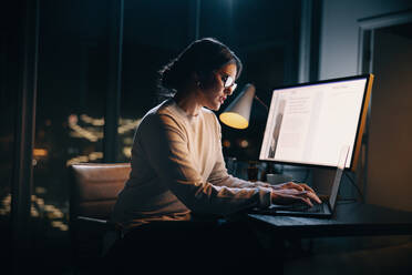 Engagierte kaukasische Geschäftsfrau, die nach Feierabend in einem schwach beleuchteten Büro arbeitet. Sie konzentriert sich auf ihren Laptop, tippt und plant. Entschlossen und professionell verkörpert sie die Vereinbarkeit von Beruf und Familie. - JLPSF30763