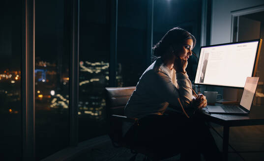 Businesswoman working late in her home office, focusing on a digital marketing project. Woman putting in long hours into her work, sitting with a laptop and a computer screen at her desk. - JLPSF30765