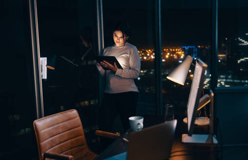 Eine entschlossene Geschäftsfrau steht in einem nächtlichen Büro, liest und analysiert mit ihrem Tablet einen Bericht. Eine professionelle Frau, die bis spät in die Nacht arbeitet, zeigt ihr Engagement für ihre Arbeit. - JLPSF30766
