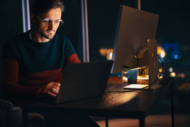 Web designer working late in an office, using his laptop to complete a project. Business man sitting at his desk, utilizing the quiet nighttime atmosphere for maximum productivity. - JLPSF30779