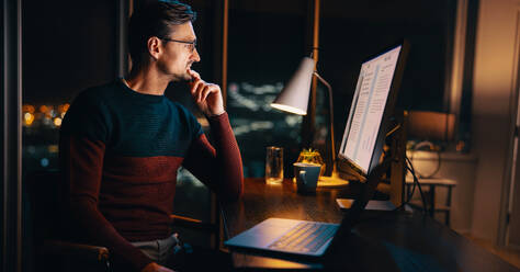 Ein weißer Geschäftsmann arbeitet bis spät in die Nacht in seinem Heimbüro an einem Marketingprojekt. Er sitzt an seinem Schreibtisch mit einem Laptop und plant und überlegt mit Hingabe. - JLPSF30783