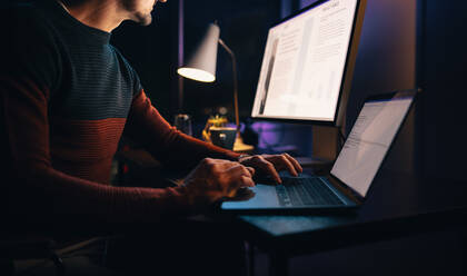 Ein weißer, männlicher Unternehmer arbeitet bis spät in die Nacht von seinem Heimbüro aus an einem Geschäftsprojekt. Er tippt auf seinem Laptop, um den Abgabetermin einzuhalten. Durch Fernarbeit beweist er Engagement und Produktivität. - JLPSF30786