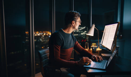 Ein zielstrebiger Geschäftsmann arbeitet bis spät in die Nacht in einem gut beleuchteten Büro an seinem Laptop. Umgeben von einem geschäftigen Arbeitsplatz arbeitet er mit Ehrgeiz und Hingabe an einem Marketingplan. - JLPSF30787