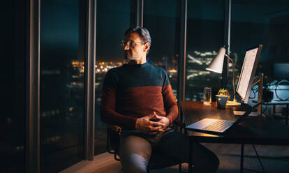 A pensive caucasian businessman working late in the office. He sits at his desk, engrossed in his laptop, looking away into the night, thinking about marketing strategies. - JLPSF30788